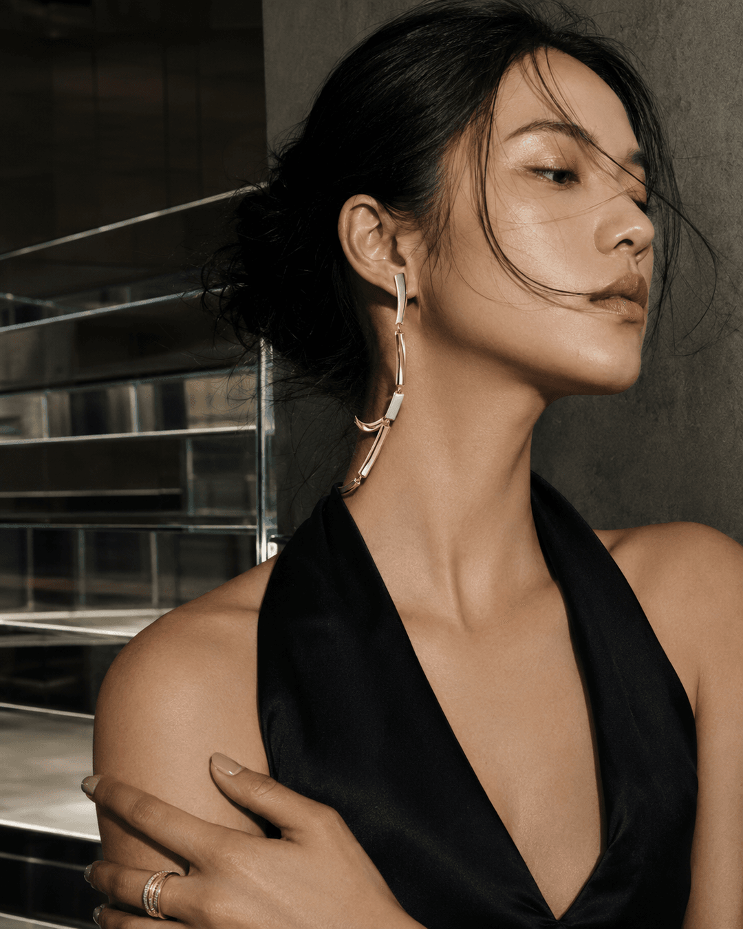 Adult East Asian model beside mirrored stairs wearing an articulated metal ear-to-collar sculpture with visible hinges and a compact ring.
