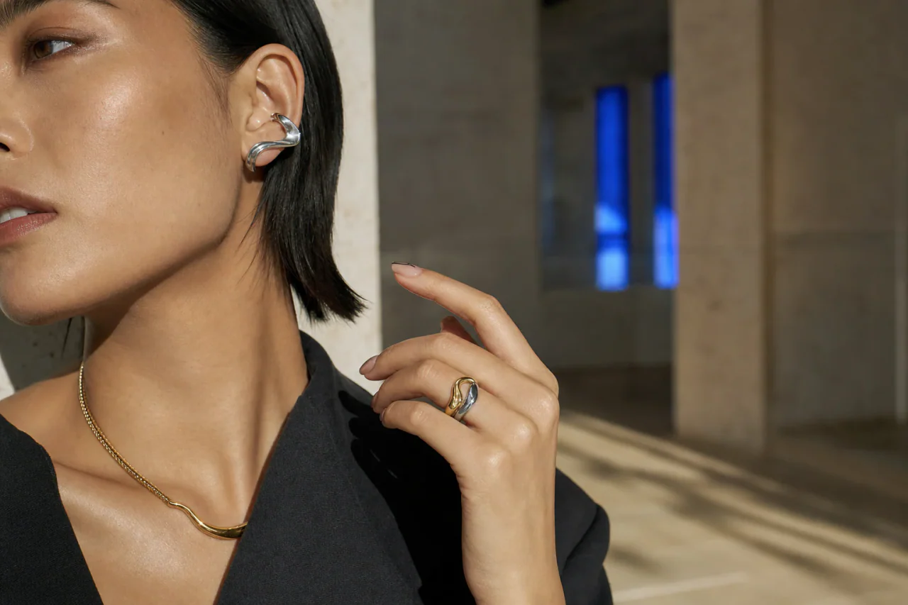 Adult model wearing a silver ear cuff, gold necklace, and mixed-tone bridge ring in a sunlit gallery corridor