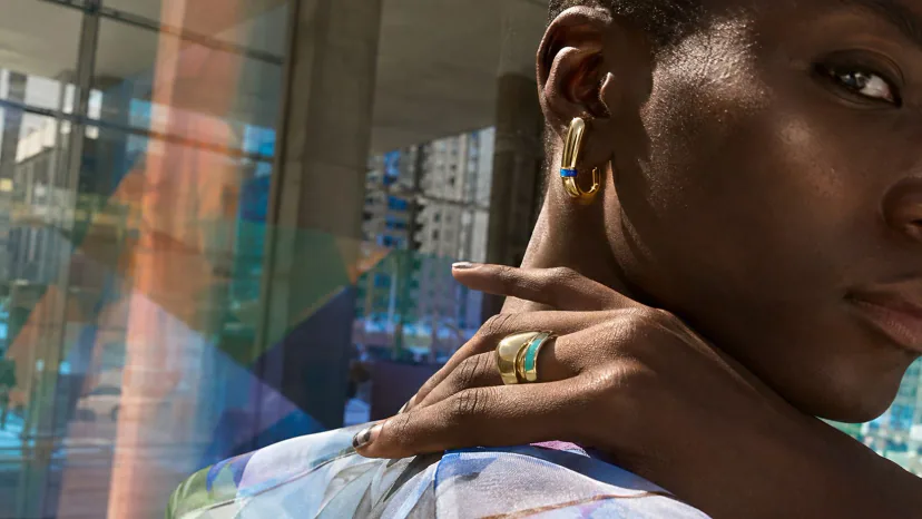 Adult Black model turning in a glass-and-concrete plaza, wearing a large polished ear sculpture and architectural ring with a narrow enamel detail.