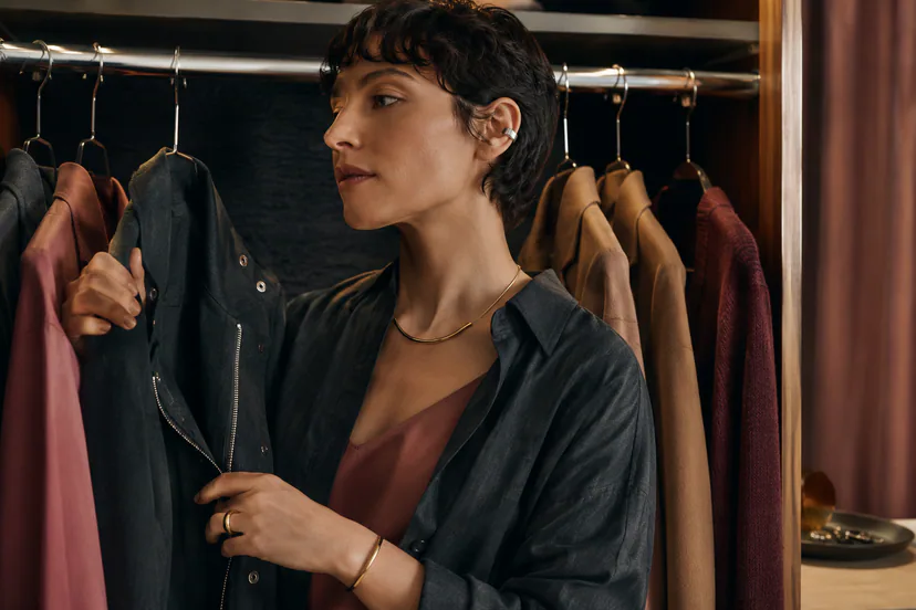 Adult wearer at an open wardrobe holds a charcoal jacket, wearing a cool-toned ear cuff and warm-toned necklace, ring, and bracelet.