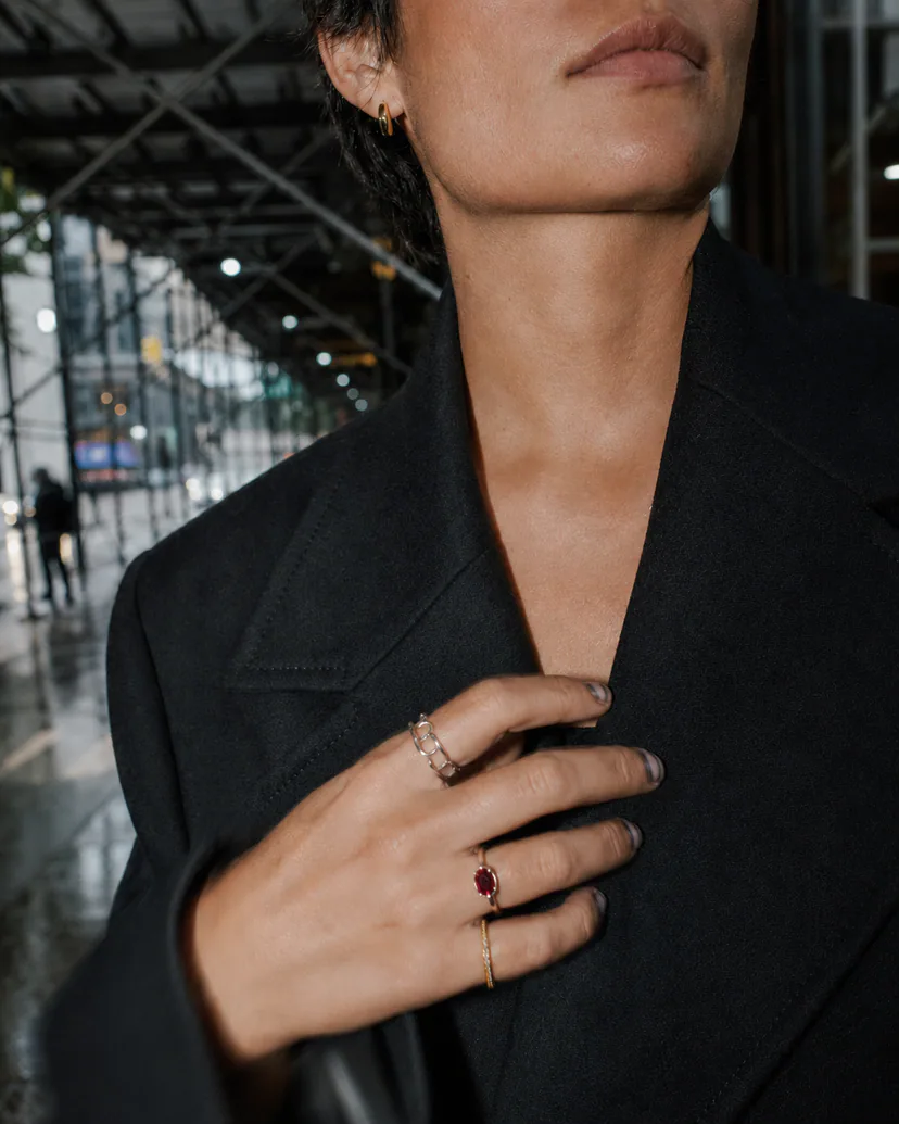 Adult model moving through an architectural passage in a black jacket with an open-link ring, red stone ring and slim gold band.