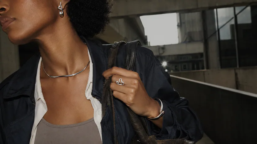 Adult Black student crossing a concrete-and-glass campus skybridge, wearing sculptural earrings, a neck piece, bracelet, and architectural ring while holding a tote.