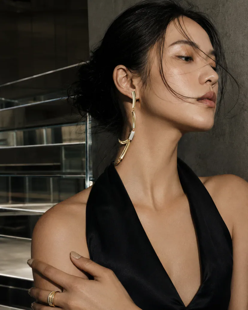 Adult East Asian model beside mirrored stairs wearing an articulated metal ear-to-collar sculpture with visible hinges and a compact ring.