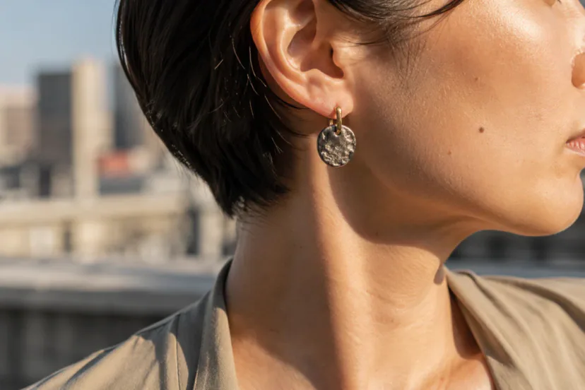 Close ear and neck view of a small textured silver earring with a warm gold bridge element in late-afternoon city light.
