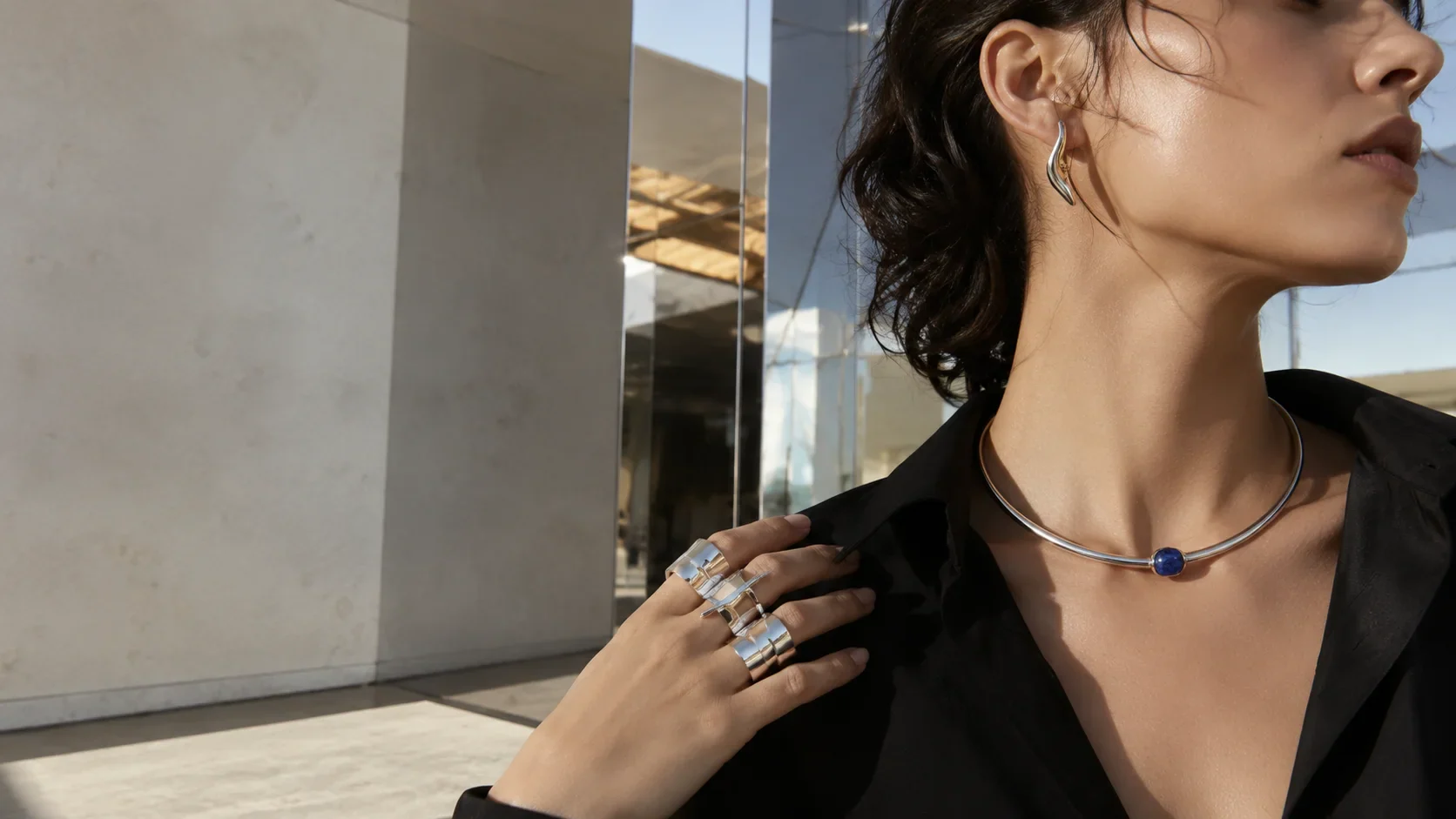 Adult wearer styling sculptural silver rings, earrings, and a collar necklace in an architectural setting