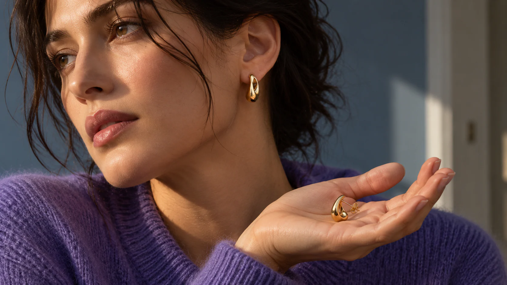 An adult in a violet knit wears a gold-colored open teardrop earring and holds its mate in an open palm.