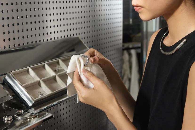 Hands gently wiping a sculptural earring before placing it in a separate fabric-lined compartment beside a ring and cuff.