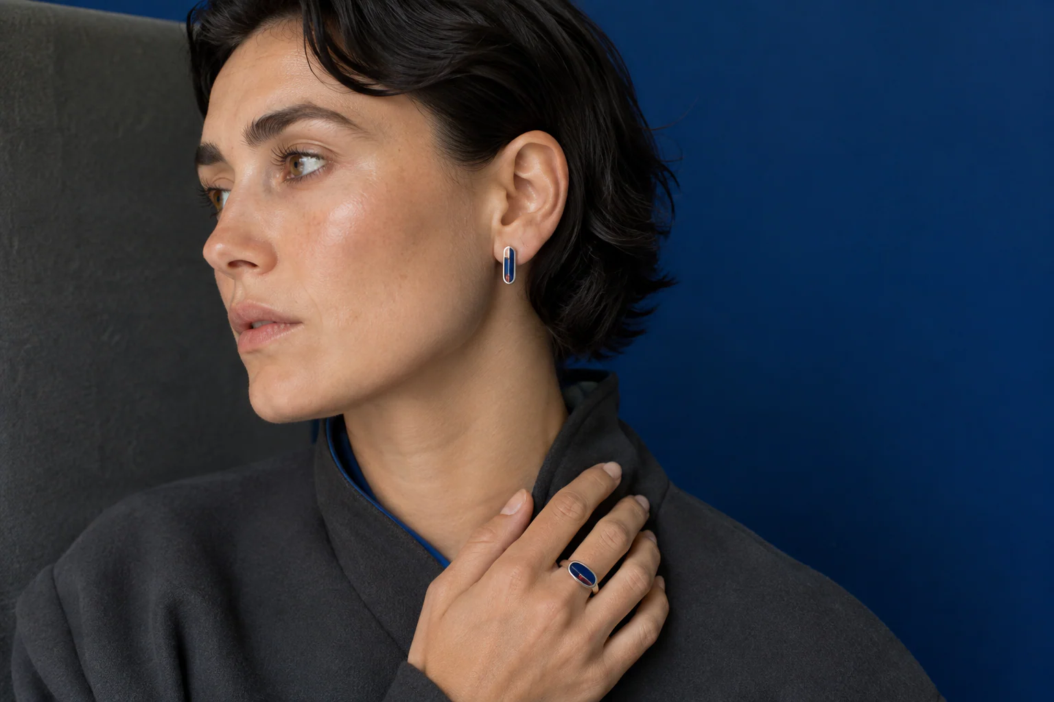 Adult in a cobalt and charcoal interior wearing a small blue enamel earring with silver edging and a compact enamel ring.