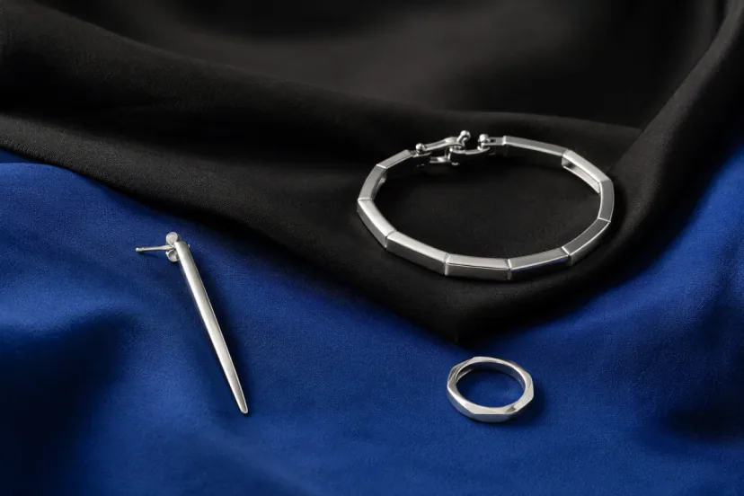 Linear silver earring, compact articulated cuff and small polished ring arranged on folded black and cobalt fabric.