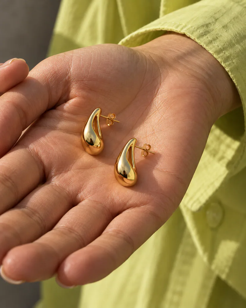 Two matching gold-colored open teardrop earrings rest in an adult’s palm beside a chartreuse shirt cuff.