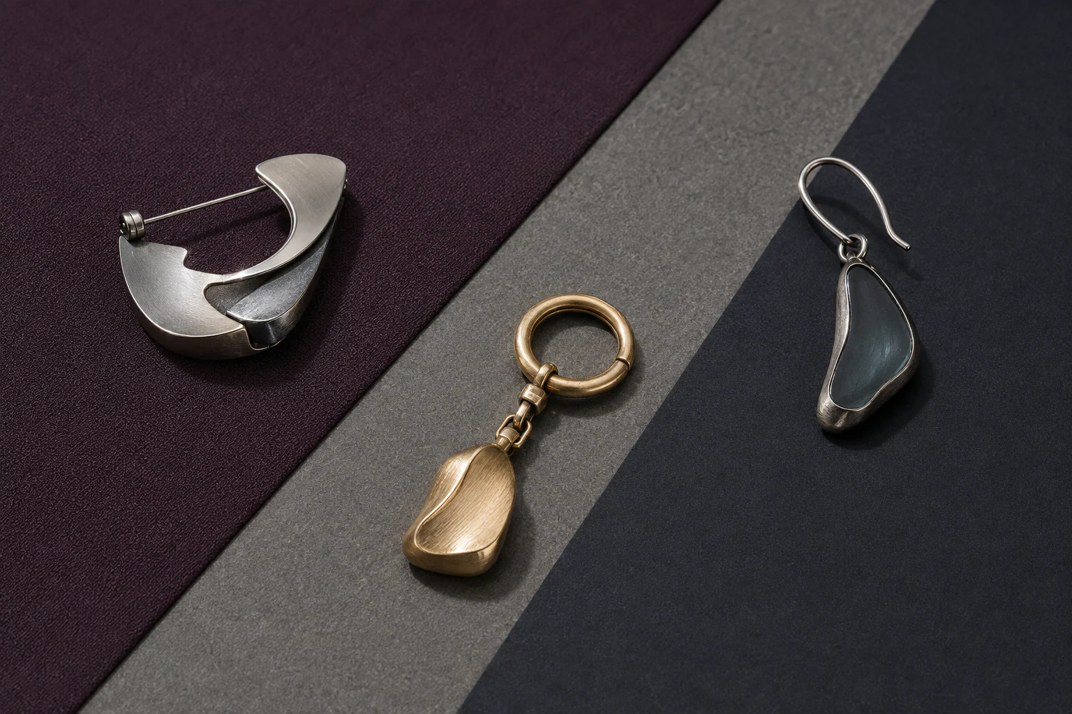 Three fictional wearable objects—a brooch, compact charm, and single asymmetric earring—arranged separately across a three-material sample board.
