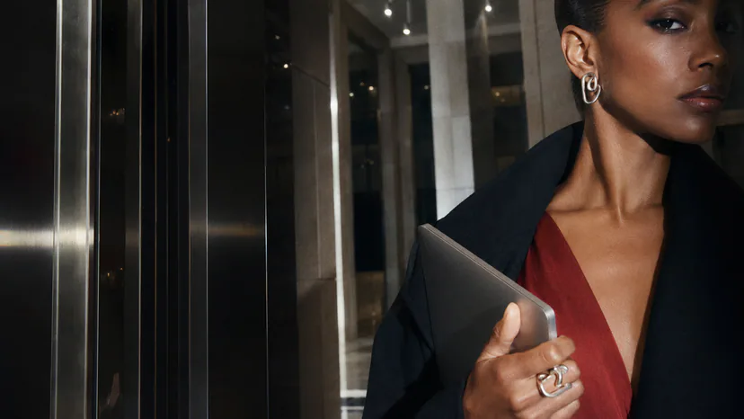 Adult Black professional in dark tailoring and a deep red top wearing linked silver earrings and a large sculptural ring while holding a notebook.