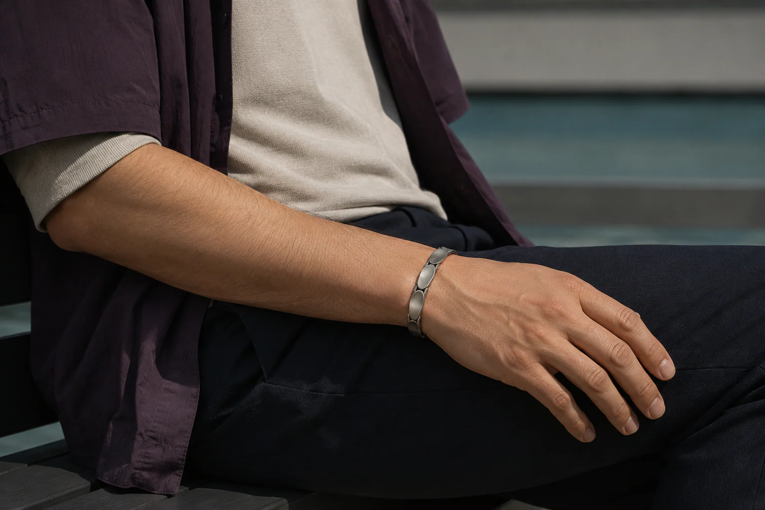 Fictional seated spectator in aubergine layers wearing one low-profile sculptural bracelet, with one forearm resting naturally across the thigh.