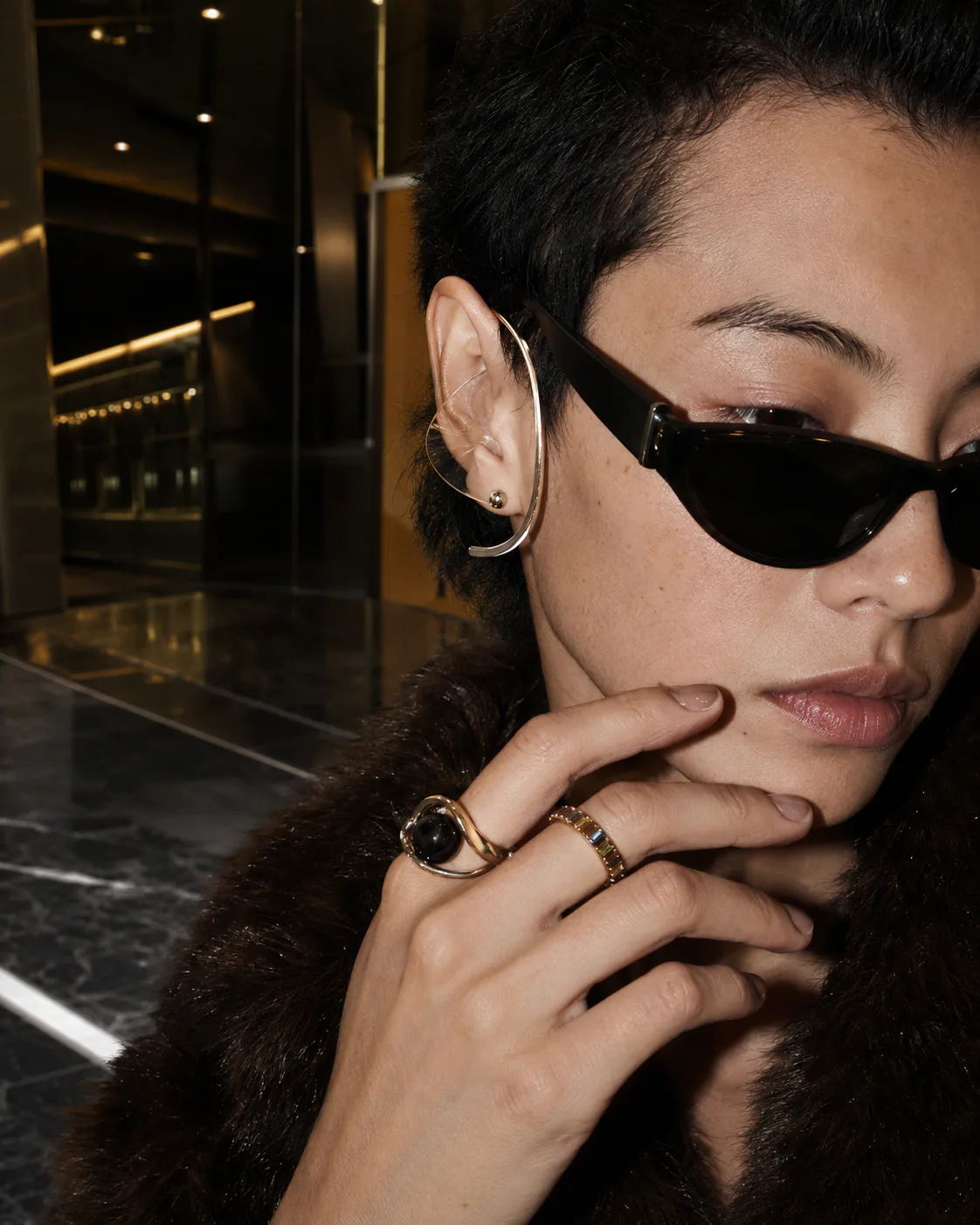 Adult East Asian model with close-cropped dark hair wearing a long sculptural silver ear piece and angular black glasses.