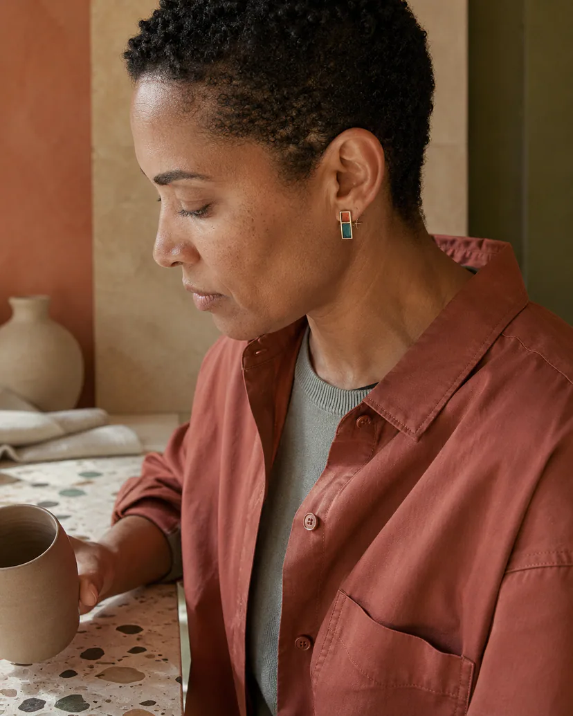 Right-profile adult in a clay-red overshirt wearing a small teal and persimmon enamel earring in a warm studio.