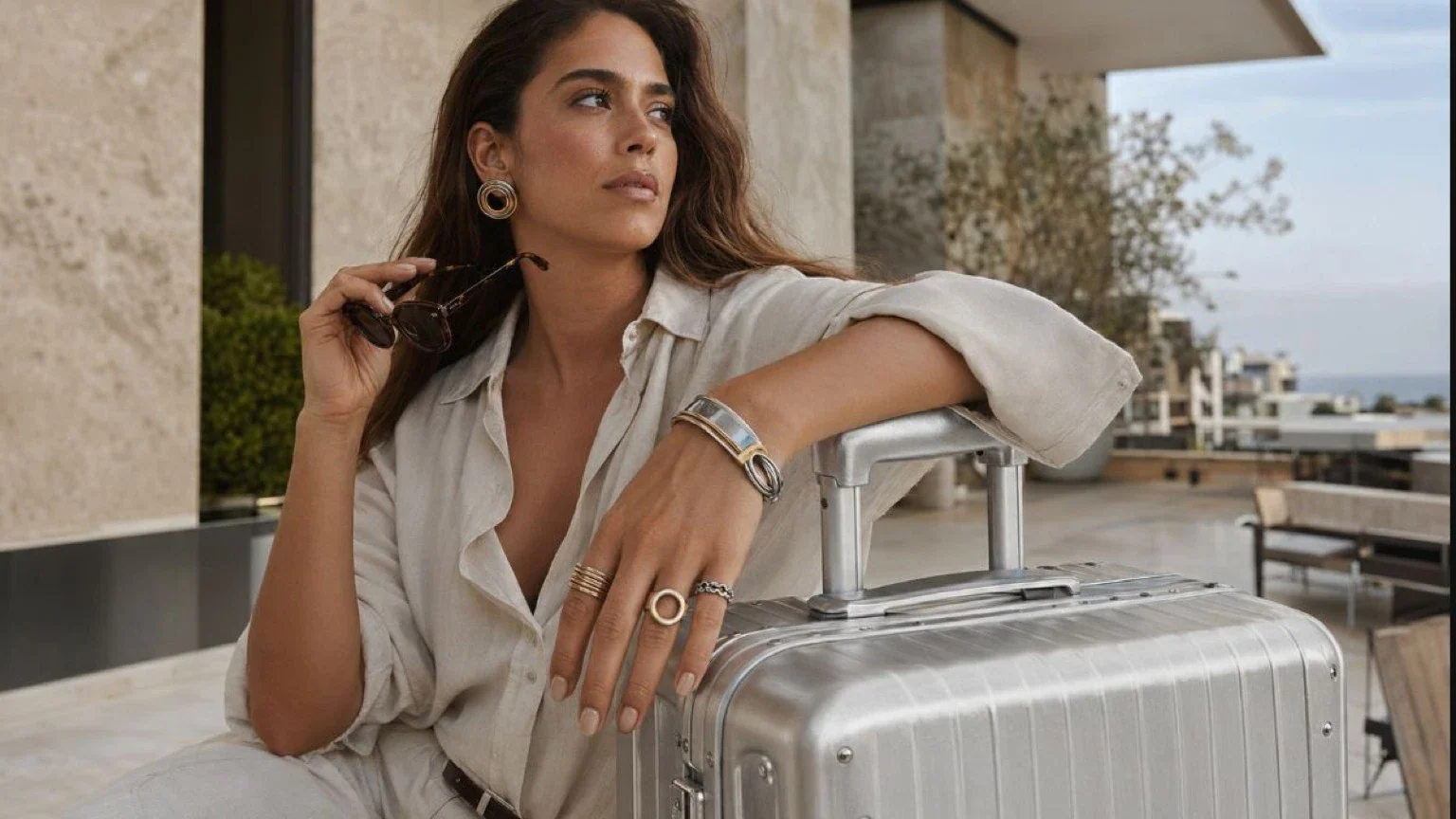 Adult traveler wearing sculptural earrings, rings and a cuff beside a silver suitcase