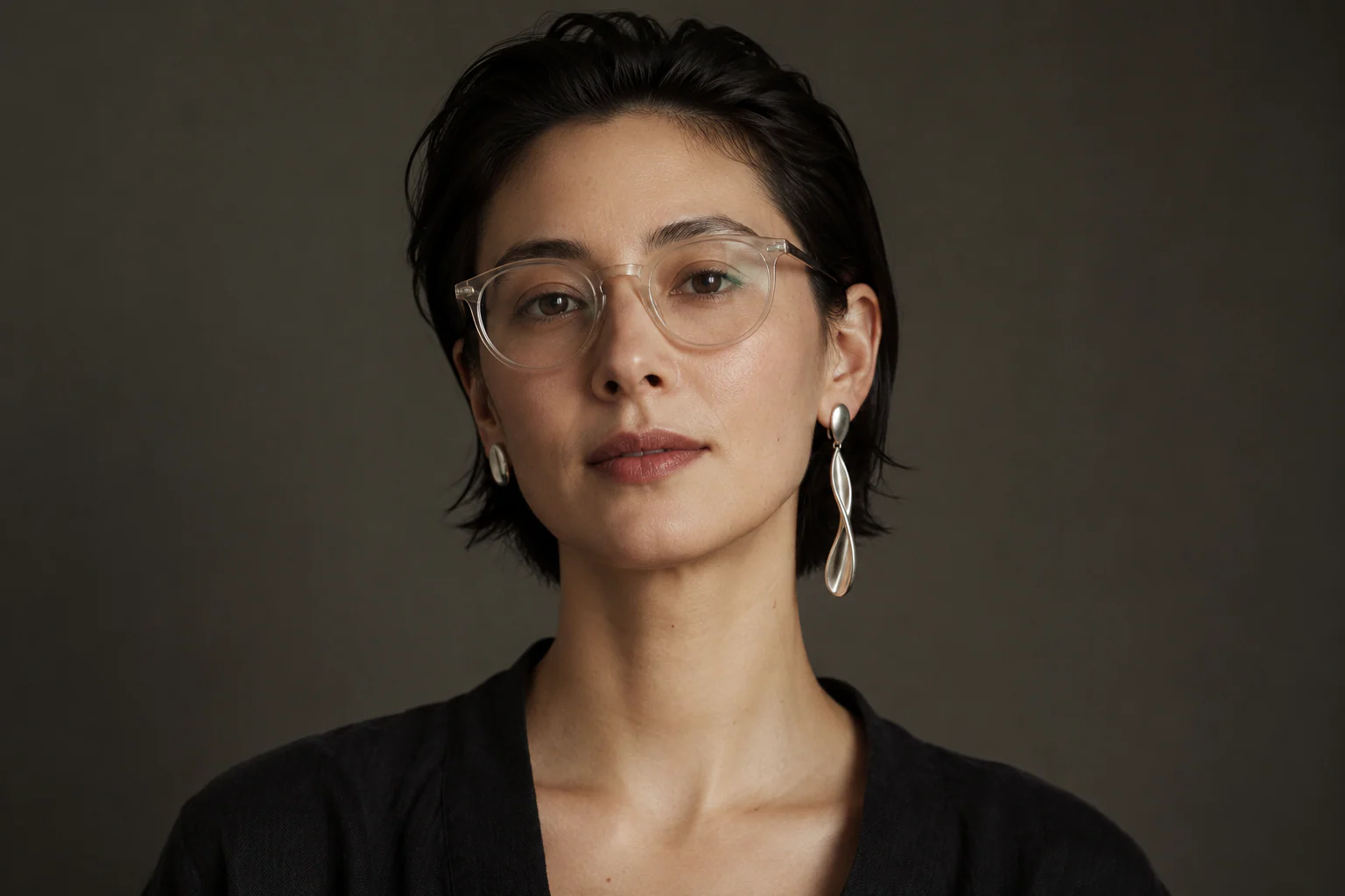 Straight-on portrait with short dark hair, translucent glasses, a long silver drop on the left ear and a compact silver oval on the right.