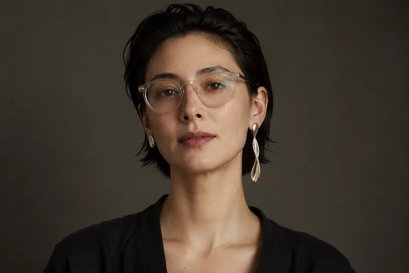 Straight-on portrait with short dark hair, translucent glasses, a long silver drop on the left ear and a compact silver oval on the right.