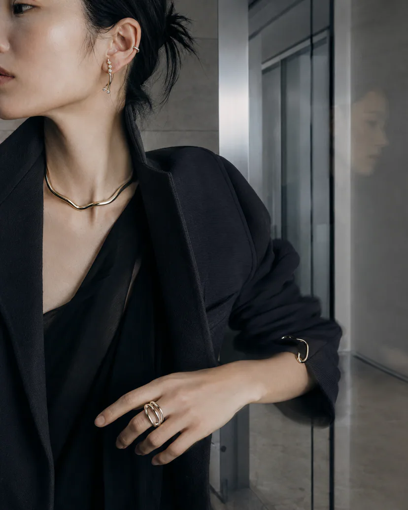 Adult East Asian professional in dark tailoring wearing a slim necklace, small ear cuff, drop earring, bracelet and sculptural ring.