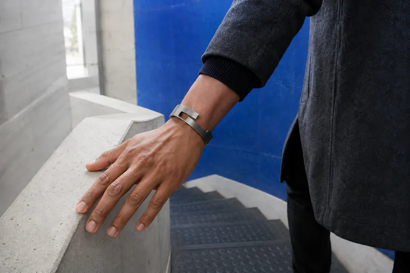 Adult left hand trails along a concrete rail while wearing one open darkened-silver cuff with a visible cut-through slot.