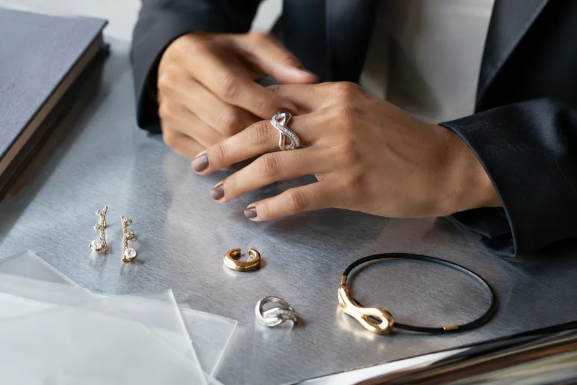 Adult professional hands at a metal desk wearing a curved silver ring beside earrings, hoops and a sculptural cord necklace.