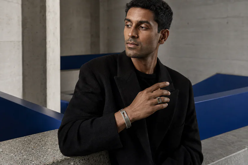Adult in black tailoring inside a concrete-and-cobalt interior, wearing a dark silver wrist cuff and an open geometric ring.