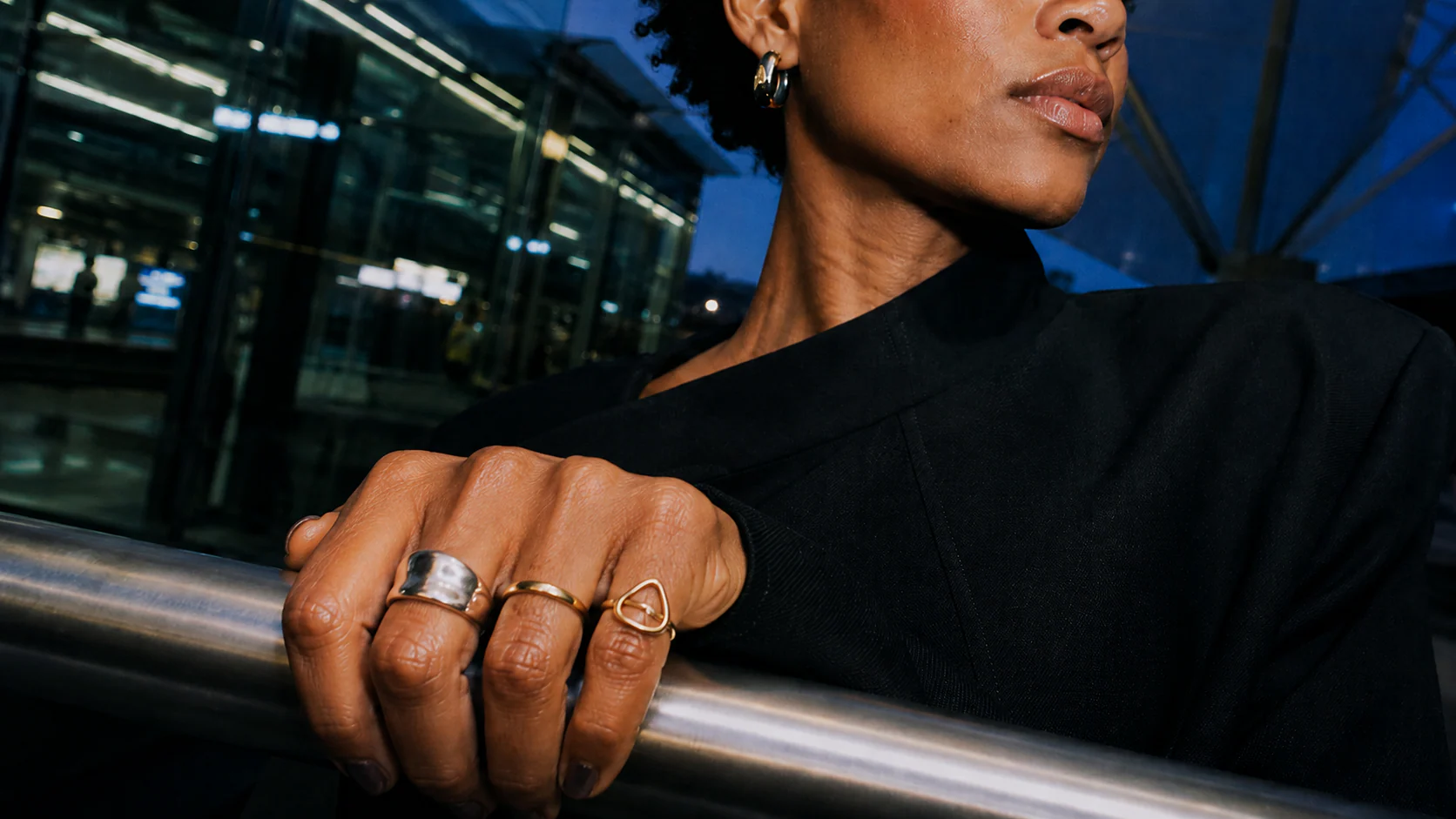 Adult Black model in an architectural transit setting wearing mixed sculptural rings and compact earrings against a black asymmetrical jacket.