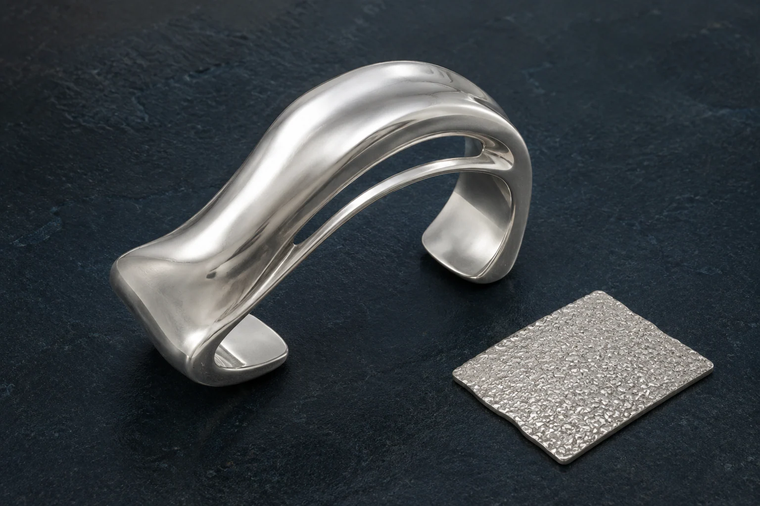 Silver sculptural open cuff with a flowing fold beside a separate granular metal texture tile on a dark mineral slab.