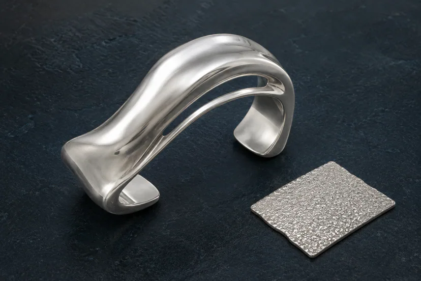 Silver sculptural open cuff with a flowing fold beside a separate granular metal texture tile on a dark mineral slab.