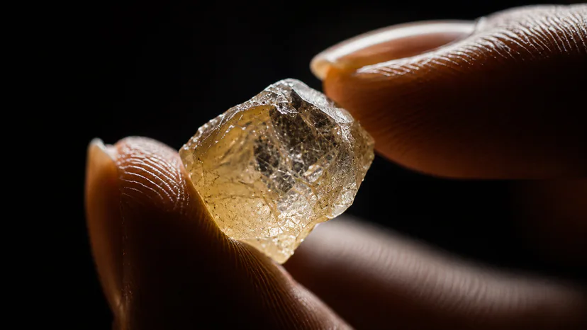 A rough pale diamond held between fingertips; appearance alone cannot prove origin or chain of custody.